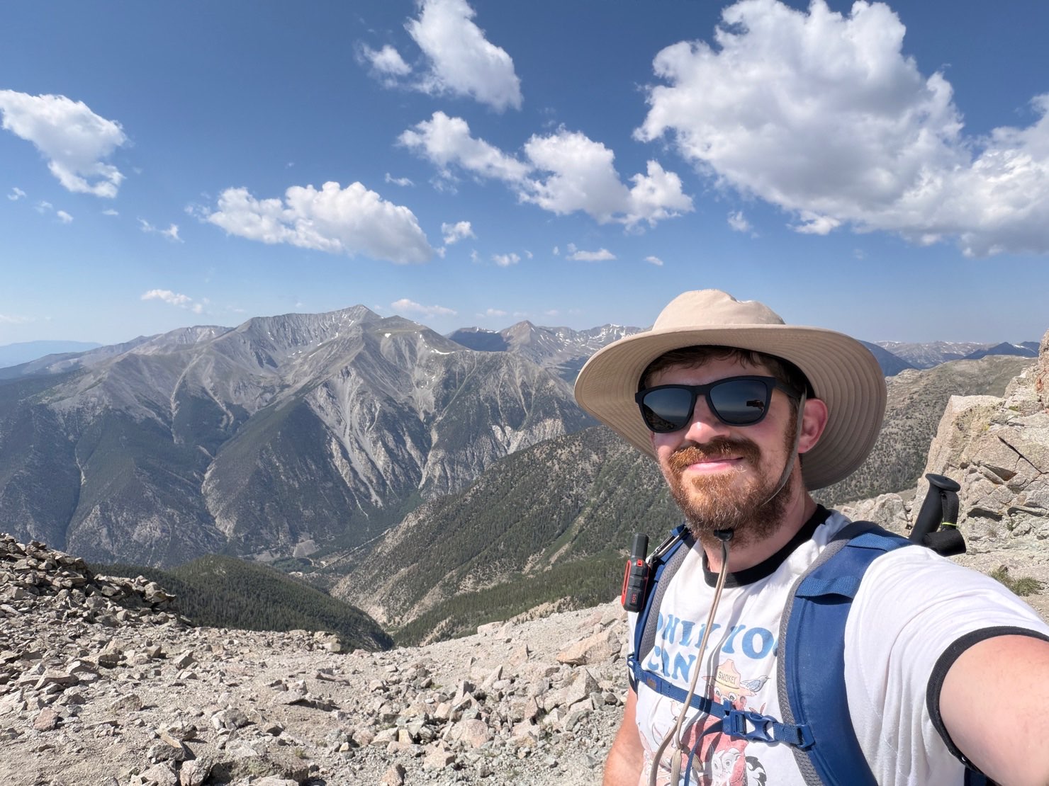 Matt on a rocky summit in a sun hat and sunglasses, with a high alpine ridgeline and a forested valley behind